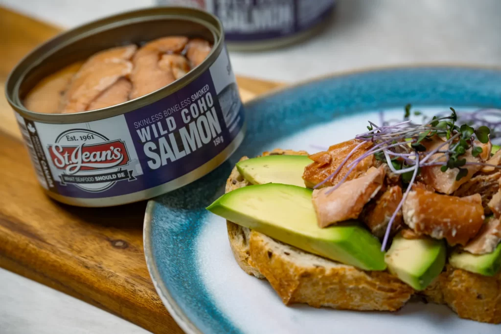 close up shot of canned wild smoked coho salmon and coho salmon on avocado toast, sitting on a blue and white plate.