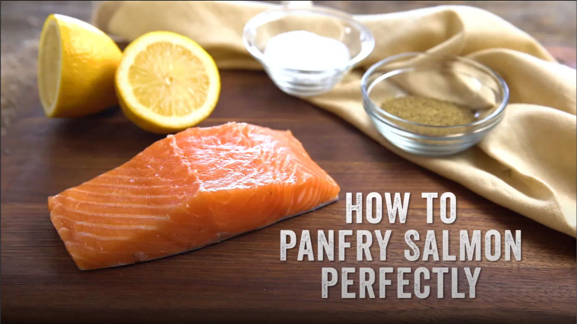 Raw salmon fillet ready to be pan-fried, laid on a table with fresh lemon slices, a clear bowl of olive oil, and salt in the background - essential ingredients for perfect West Coast style pan-fried salmon.