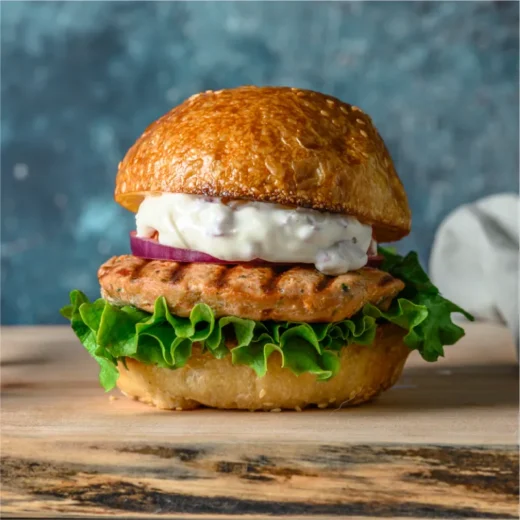 Image depicts a 'Wild Salmon Grill Master Box' featuring a grilled salmon burger nestled in an artisan bun. Bright green lettuce, thinly sliced red onions, and a glossy layer of mayo sauce are visible within the burger, indicating a mix of textures and flavors.